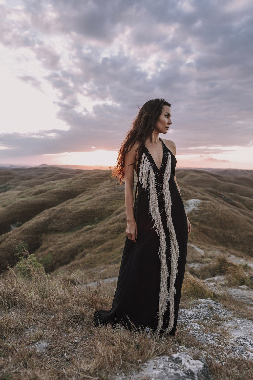 Clad in the Black Cotton Long Maxi Dress • Open Back Goddess Dress by AYA Sacred Wear, a woman stands gracefully on a grassy hilltop at sunset. She gazes into the distance, surrounded by rolling hills and a striking, cloudy sky in the backdrop.