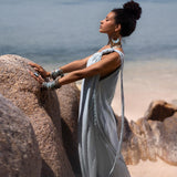 A woman clad in an elegant Blue Aurora Greek Goddess Dress by AYA Sacred Wear, adorned with various accessories, stands atop large rocks overlooking a serene beach and ocean. Her arms and upper body are slightly tilted back, eyes closed, as she appears to be enjoying the tranquil scenery.