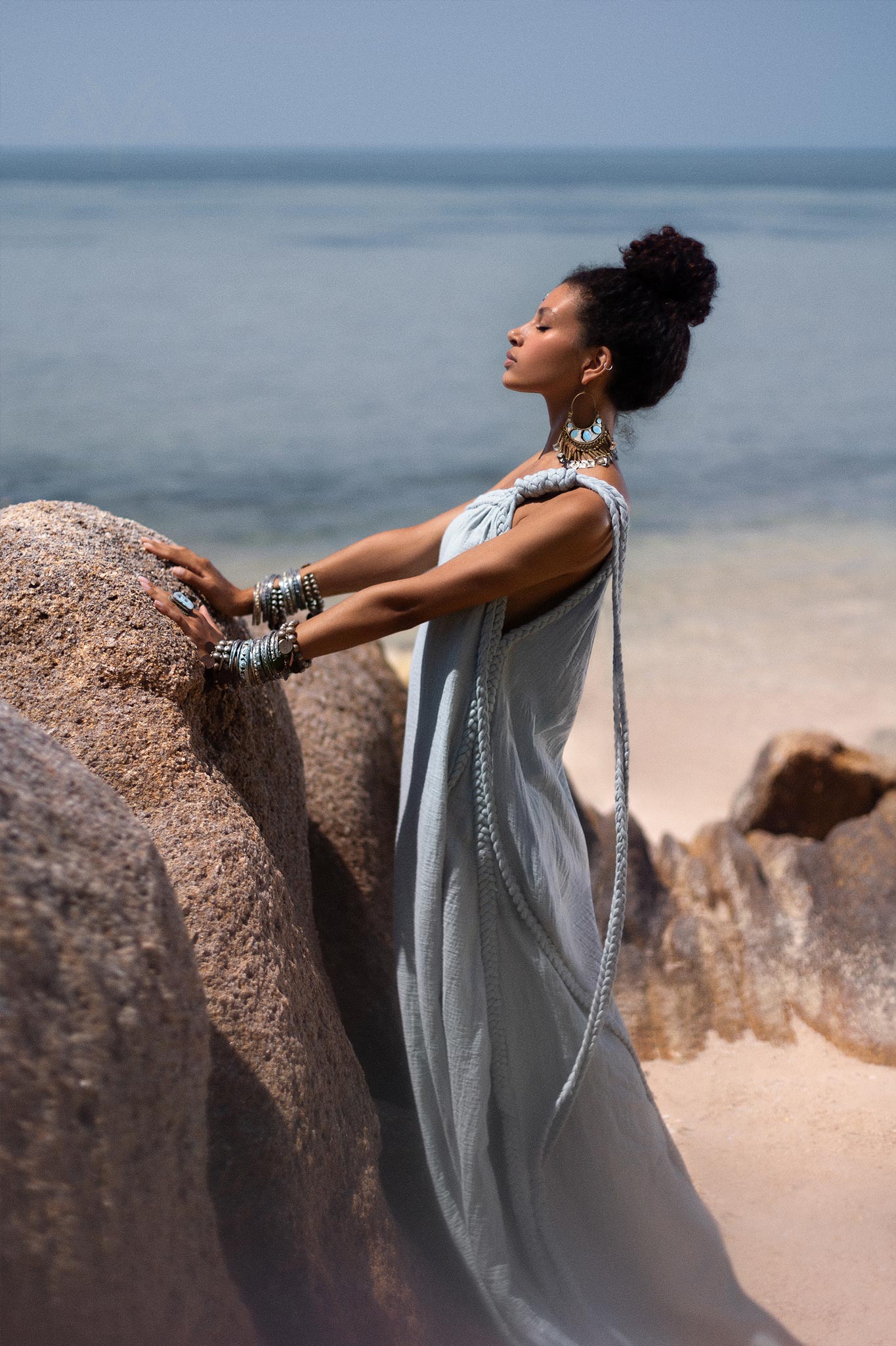A woman clad in an elegant Blue Aurora Greek Goddess Dress by AYA Sacred Wear, adorned with various accessories, stands atop large rocks overlooking a serene beach and ocean. Her arms and upper body are slightly tilted back, eyes closed, as she appears to be enjoying the tranquil scenery.