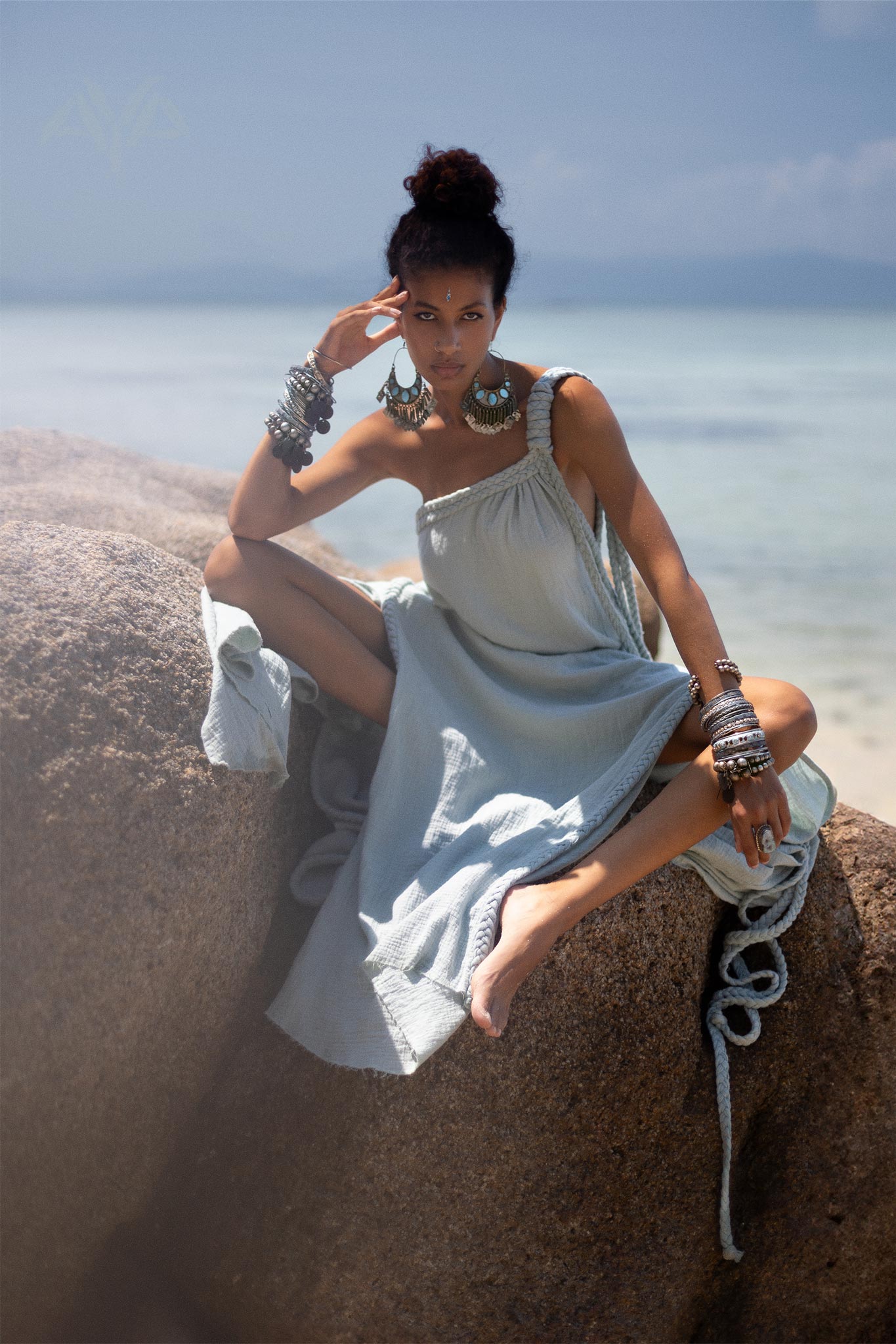 A person with dark hair is sitting on a large rock on a sandy beach, wearing an elegant Blue Aurora Greek Goddess Dress by AYA Sacred Wear. The Macrame Toga Dress, with its one-shoulder maxi design, is made of organic cotton. They are adorned with large, bold bracelets and earrings. The serene blue ocean and distant horizon provide a tranquil backdrop.
