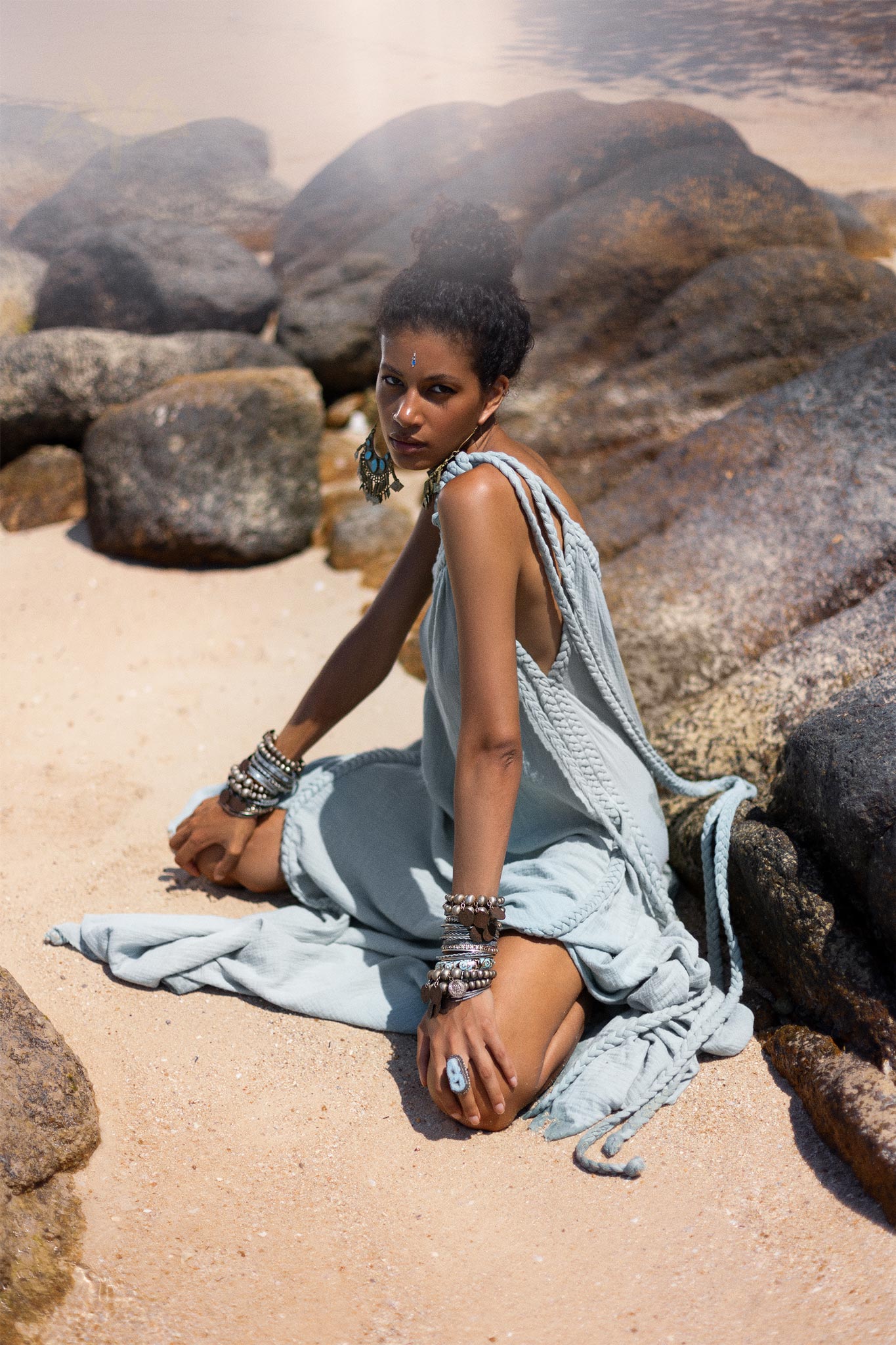 A woman with short, curly hair sits on a sandy beach surrounded by large rocks. She wears a flowing Blue Aurora Macrame Toga Dress, also known as the One Shoulder Maxi Dress from AYA Sacred Wear, adorned with multiple bracelets and earrings. She looks towards the camera with a soft, contemplative expression as the sun casts a warm glow on the serene scene.