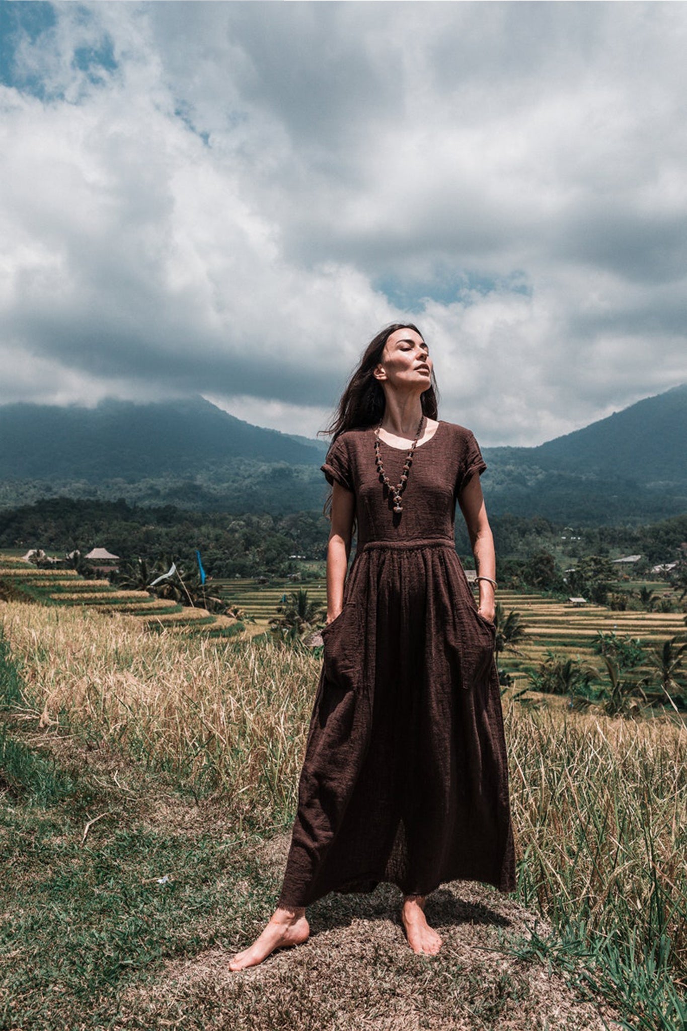 In a lush, grassy field with mountains looming in the background, a woman stands barefoot wearing an AYA Sacred Wear Bohemian Boho Dress for Women. The handwoven organic cotton fabric flows gracefully as she gazes upwards with a serene expression, embodying the essence of sustainable ethical fashion under a partly cloudy sky.
