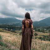 A woman stands on a grassy hill with her back to us, dressed in an AYA Sacred Wear Bohemian Boho Dress for Women. She gazes over verdant rice paddies and a mountainous view beneath a partly cloudy sky, truly representing the essence of sustainable ethical fashion.