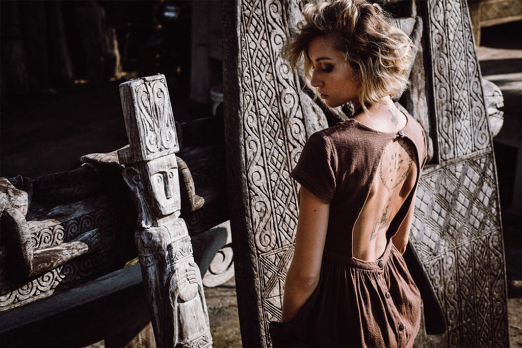 A woman with short hair wearing an AYA Sacred Wear Bohemian Boho Dress for Women stands alongside intricately carved wooden statues, her upper back displaying a tattoo. Natural light illuminates the detailed patterns on both the statues and her dress.