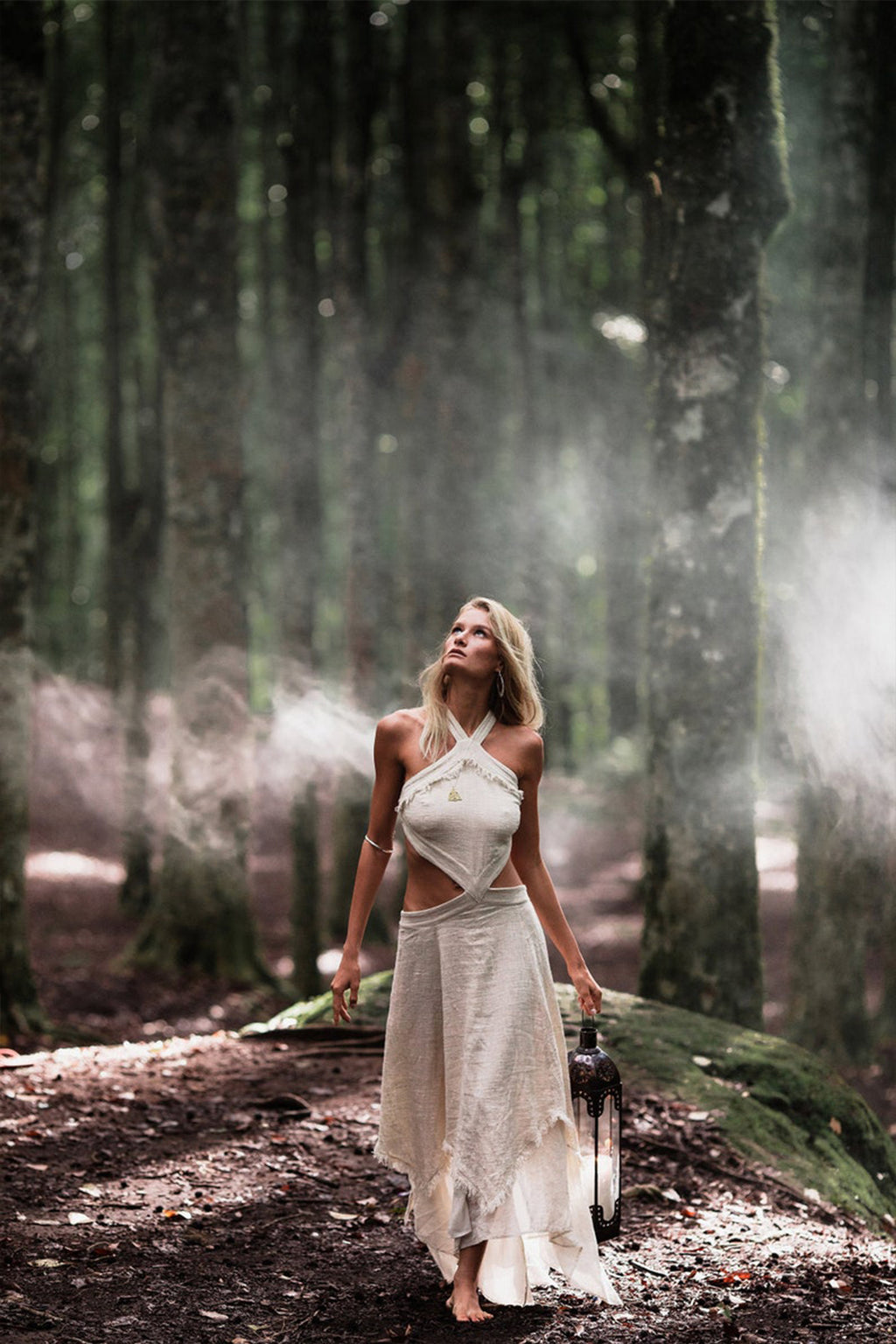 A woman in an elegant Bohemian Boho Wedding Dress from AYA Sacred Wear strolls barefoot through a misty forest, holding a lantern. Sunlight filters through the trees, casting a mystical glow. She gazes upward, enveloped by towering trunks and gentle light.