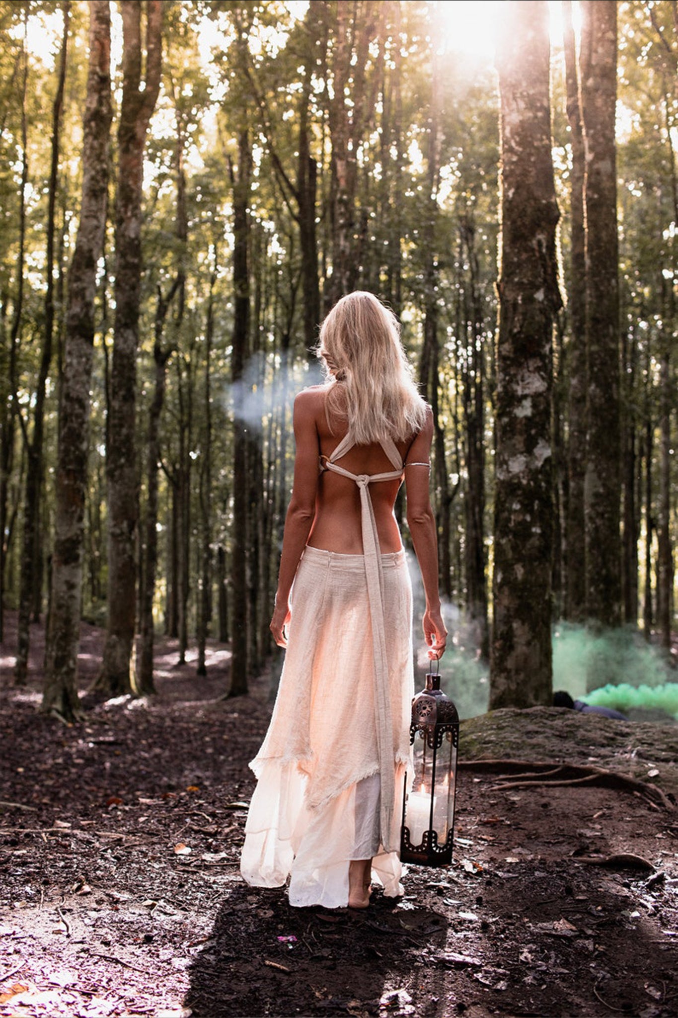 A woman with long blonde hair, wearing the Bohemian Boho Wedding Dress by AYA Sacred Wear, wanders through a sunlit forest holding a lantern. A green mist is visible among the trees in the background, creating a serene and ethereal scene.