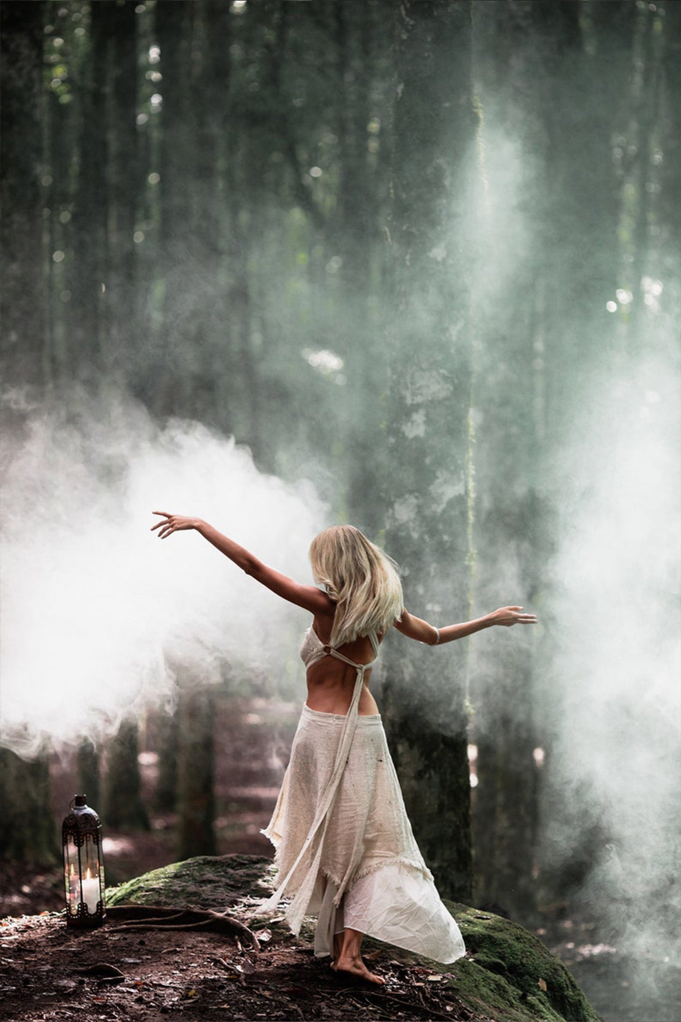 A woman wearing the Bohemian Boho Wedding Dress from AYA Sacred Wear dances along a misty forest path, flanked by towering trees. A lantern at her side casts an ethereal glow over the enchanting scene.