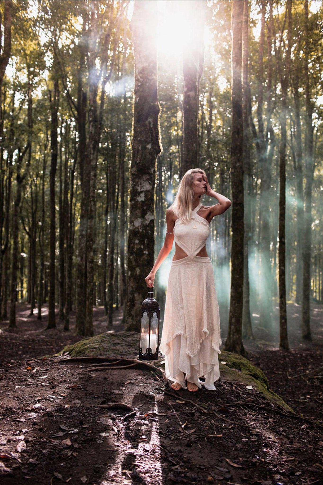 A woman wearing the Bohemian Boho Wedding Dress by AYA Sacred Wear stands barefoot in a forest, holding a lantern. Sunlight filters through the trees, casting beams of light around her. She gazes upward, her other hand resting on her head.