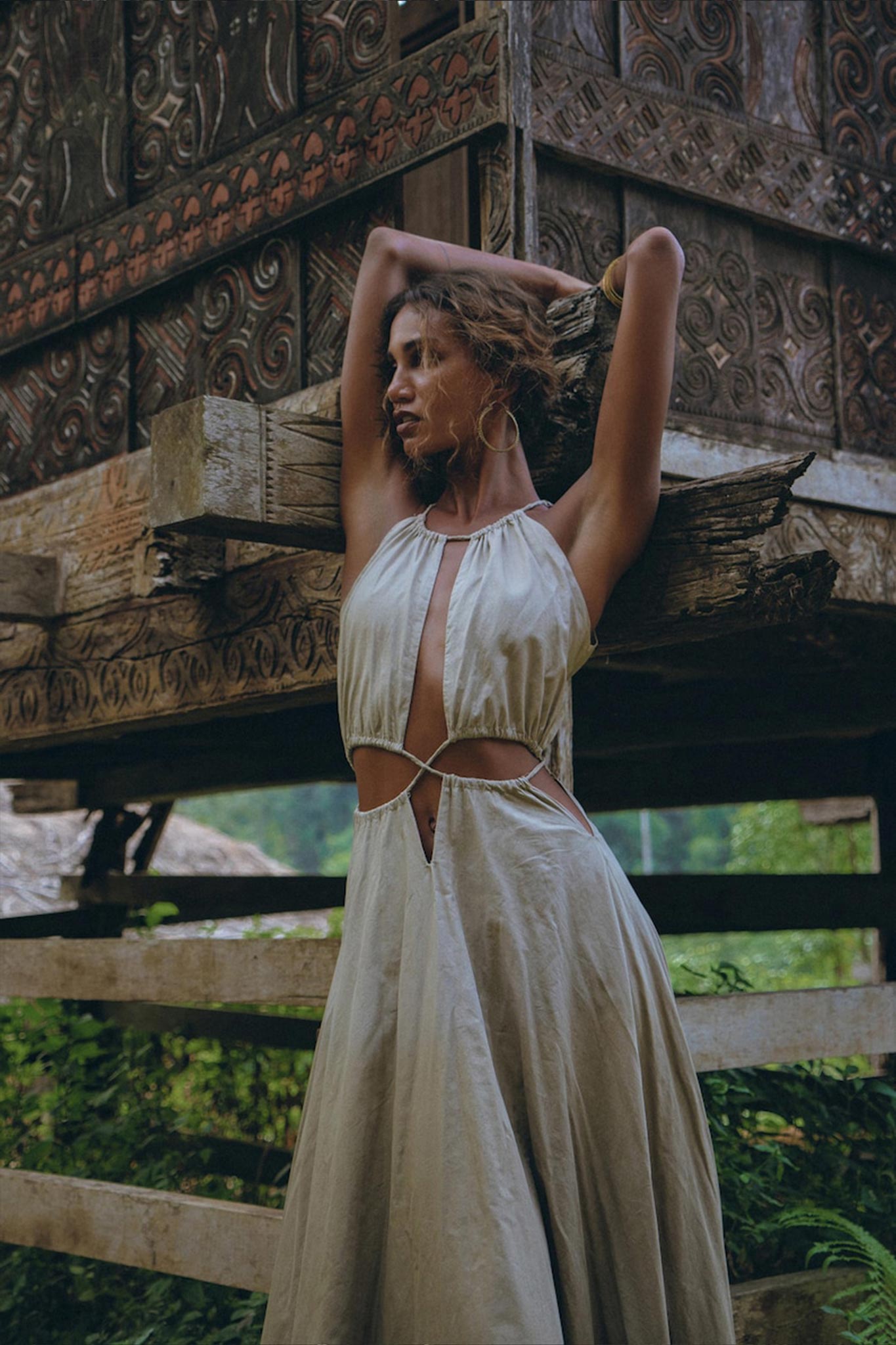 A person in a sleeveless, light-gray bohemian dress from AYA Sacred Wear stands with arms raised against a rustic, carved wooden structure. The Boho Bridesmaid Dress for Women, with its open back and cut-out sides, is crafted from organic fabric. Lush greenery can be seen in the background.