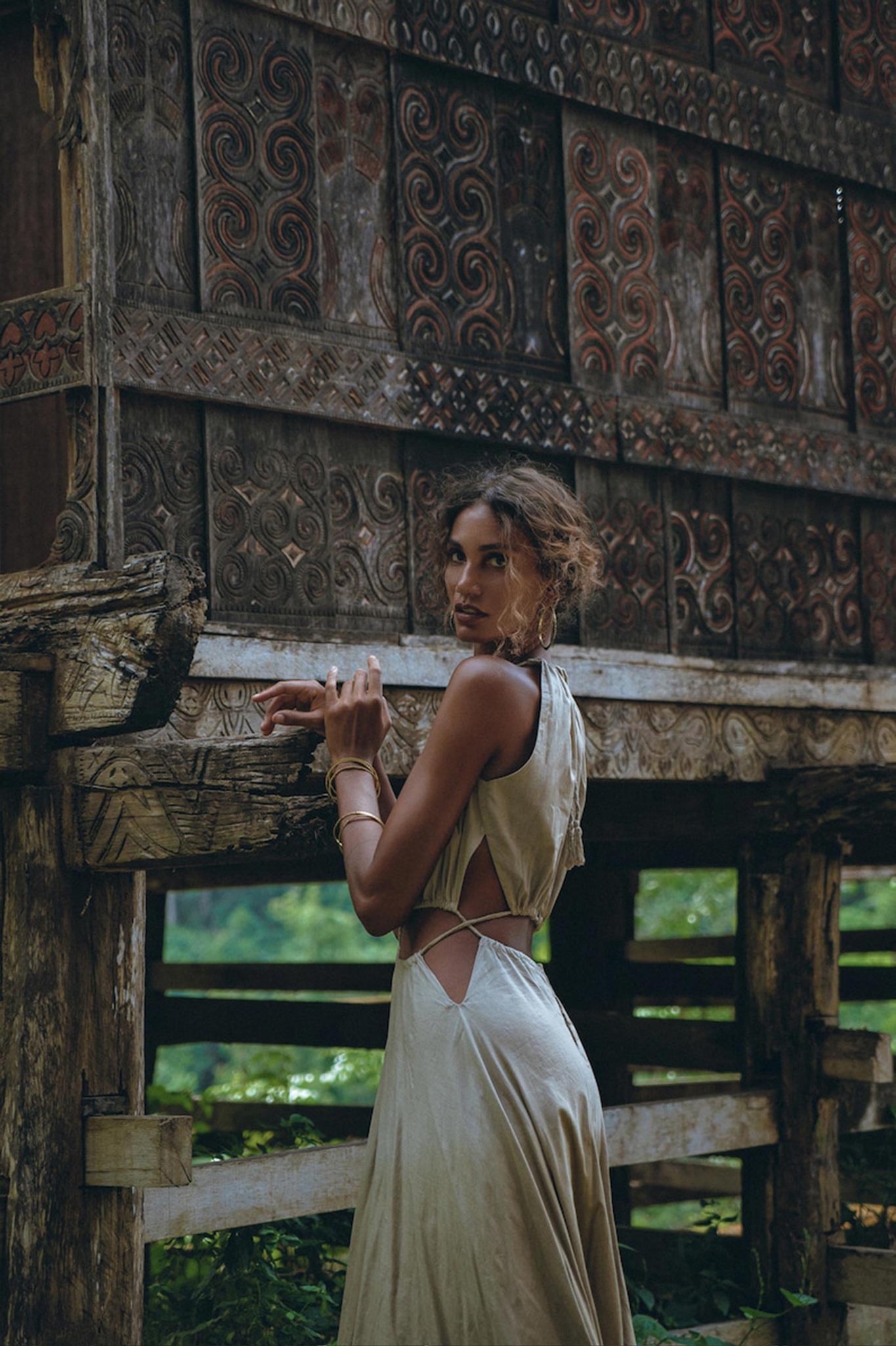 A person in an AYA Sacred Wear boho bridesmaid dress for women, featuring open back sides and a belly cutout, stands beside an ornately carved wooden structure with intricate patterns. The lush green foliage in the background complements the organic fabric and enhances the serene, natural ambiance.