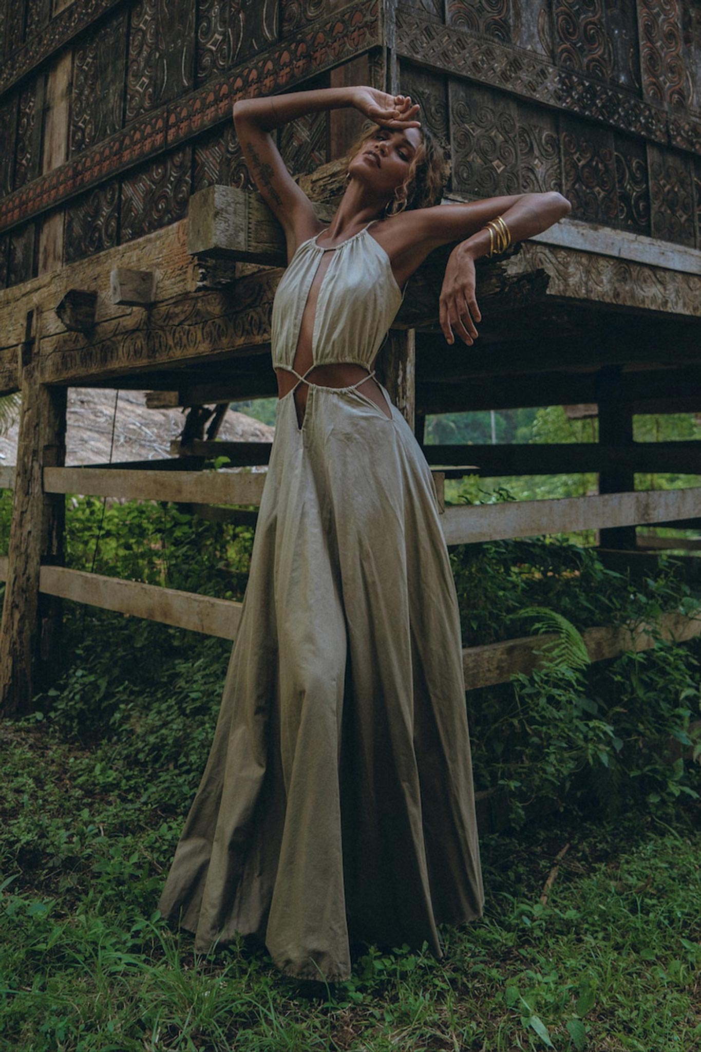 A person poses outdoors in a long, flowing boho bridesmaid dress with open back and side details by AYA Sacred Wear. The background features a rustic wooden structure with intricate carvings. Greenery surrounds the area, enhancing the natural, serene setting and perfectly complementing the organic fabric.