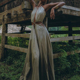 A person poses outdoors in a long, flowing boho bridesmaid dress with open back and side details by AYA Sacred Wear. The background features a rustic wooden structure with intricate carvings. Greenery surrounds the area, enhancing the natural, serene setting and perfectly complementing the organic fabric.