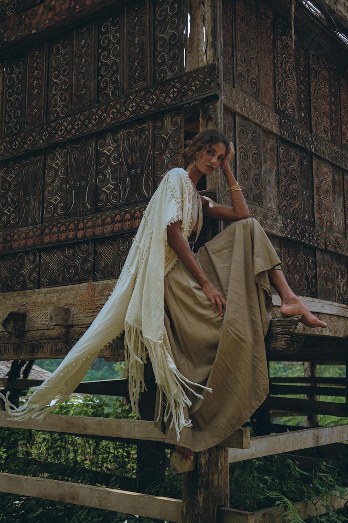 A person is sitting on a wooden structure adorned with intricate carvings, wearing a beige Boho Bridesmaid Dress from AYA Sacred Wear. The open back and sides of the dress create a harmonious blend with the natural outdoor setting, surrounded by lush greenery and exuding a rustic charm.