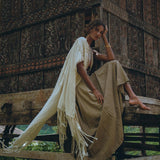 A person is sitting on a wooden structure adorned with intricate carvings, wearing a beige Boho Bridesmaid Dress from AYA Sacred Wear. The open back and sides of the dress create a harmonious blend with the natural outdoor setting, surrounded by lush greenery and exuding a rustic charm.