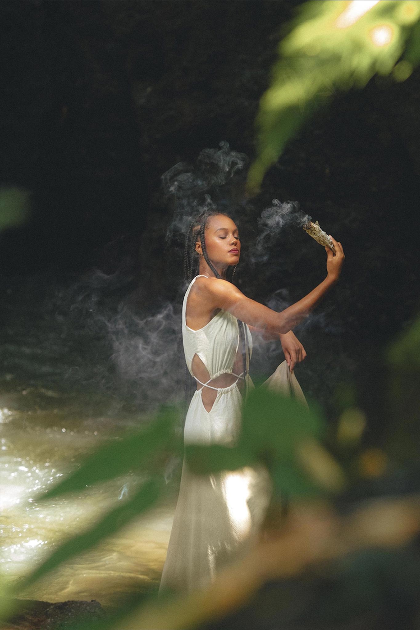 A woman in a flowing Boho Bridesmaid Dress for Women from AYA Sacred Wear stands near water, holding a smoking incense stick. Surrounded by greenery, she appears calm and immersed in a natural setting, with sunlight filtering through leaves.