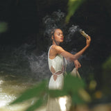 A woman in a flowing Boho Bridesmaid Dress for Women from AYA Sacred Wear stands near water, holding a smoking incense stick. Surrounded by greenery, she appears calm and immersed in a natural setting, with sunlight filtering through leaves.