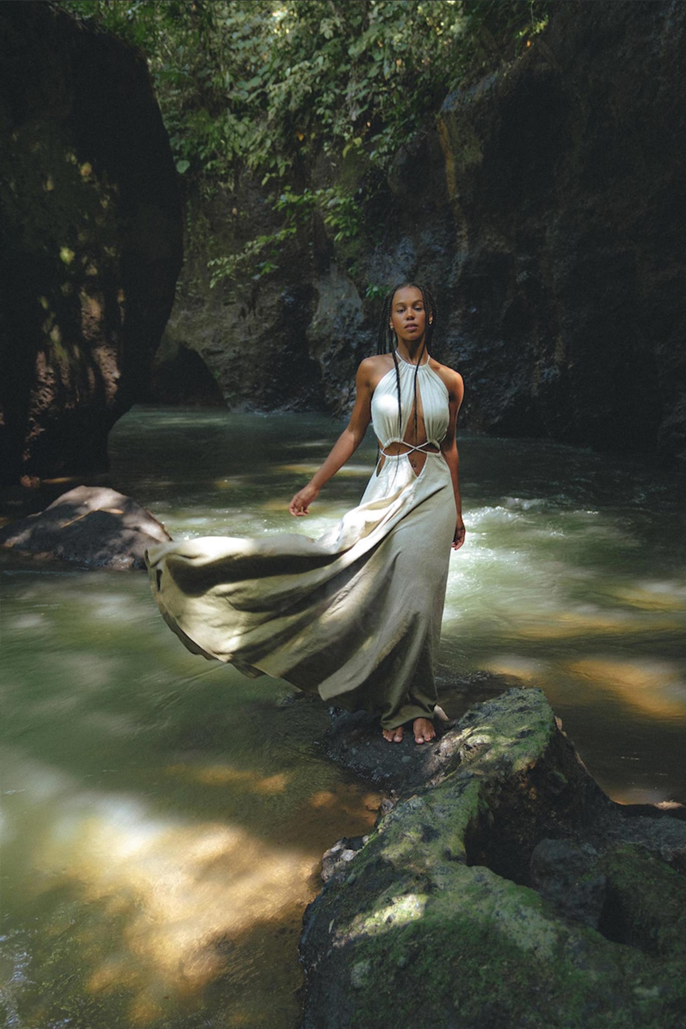 A woman wearing a Boho Bridesmaid Dress for Women by AYA Sacred Wear stands on a moss-covered rock beside a stream in the forest. Sunlight filters through the trees, creating a peaceful, natural setting that perfectly complements her open-back organic fabric dress.