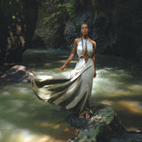 A woman wearing a Boho Bridesmaid Dress for Women by AYA Sacred Wear stands on a moss-covered rock beside a stream in the forest. Sunlight filters through the trees, creating a peaceful, natural setting that perfectly complements her open-back organic fabric dress.