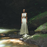 A woman in an AYA Sacred Wear Boho Bridesmaid Dress for Women, with its open back sides and flowing organic fabric, stands gracefully on a rock beside a tranquil stream. Sunlight filters through the trees, casting dappled shadows on the lush greenery around her, creating an ambiance that perfectly evokes the serenity of Ayurveda.