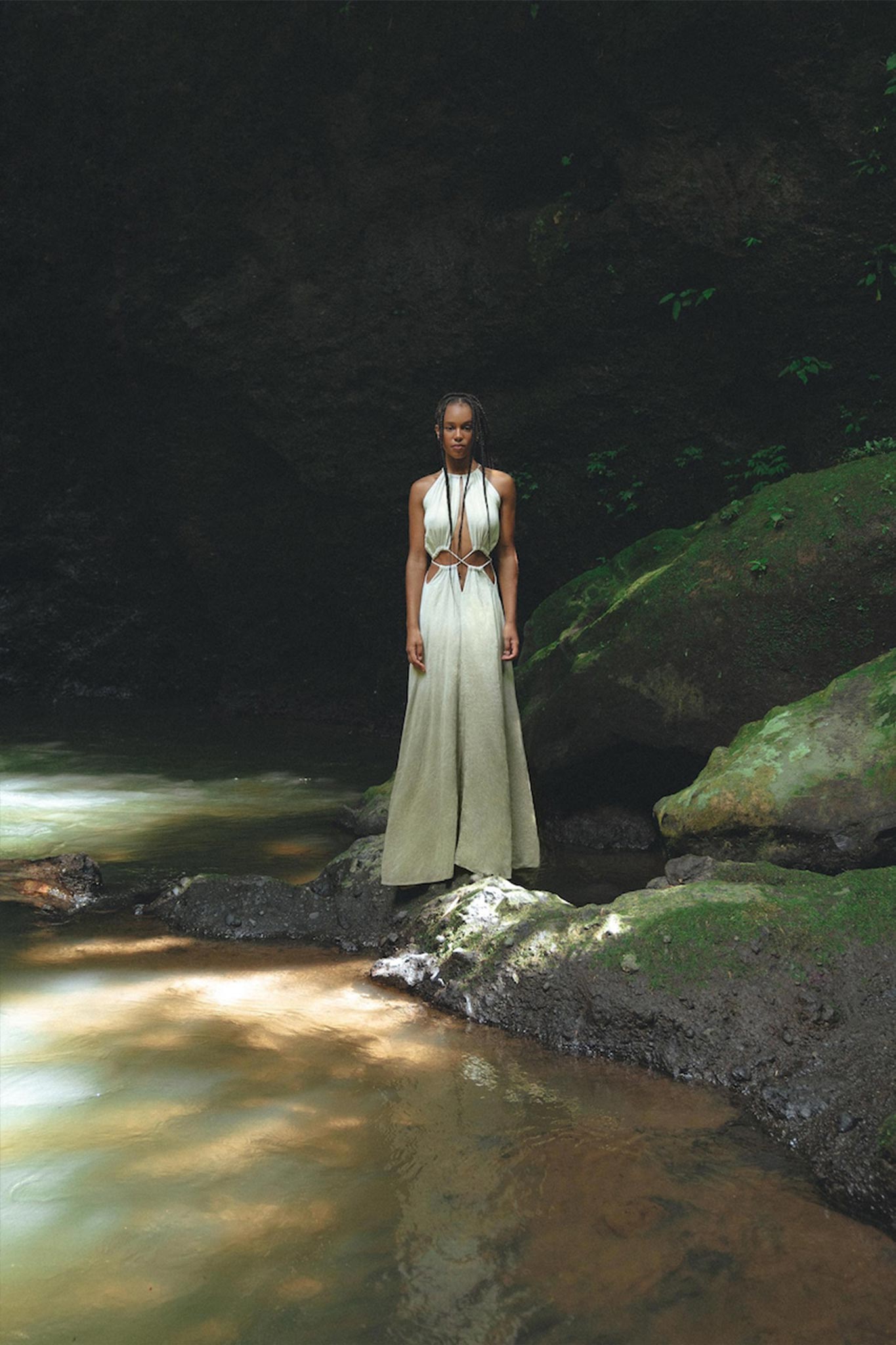 A woman in an AYA Sacred Wear Boho Bridesmaid Dress for Women, with its open back sides and flowing organic fabric, stands gracefully on a rock beside a tranquil stream. Sunlight filters through the trees, casting dappled shadows on the lush greenery around her, creating an ambiance that perfectly evokes the serenity of Ayurveda.