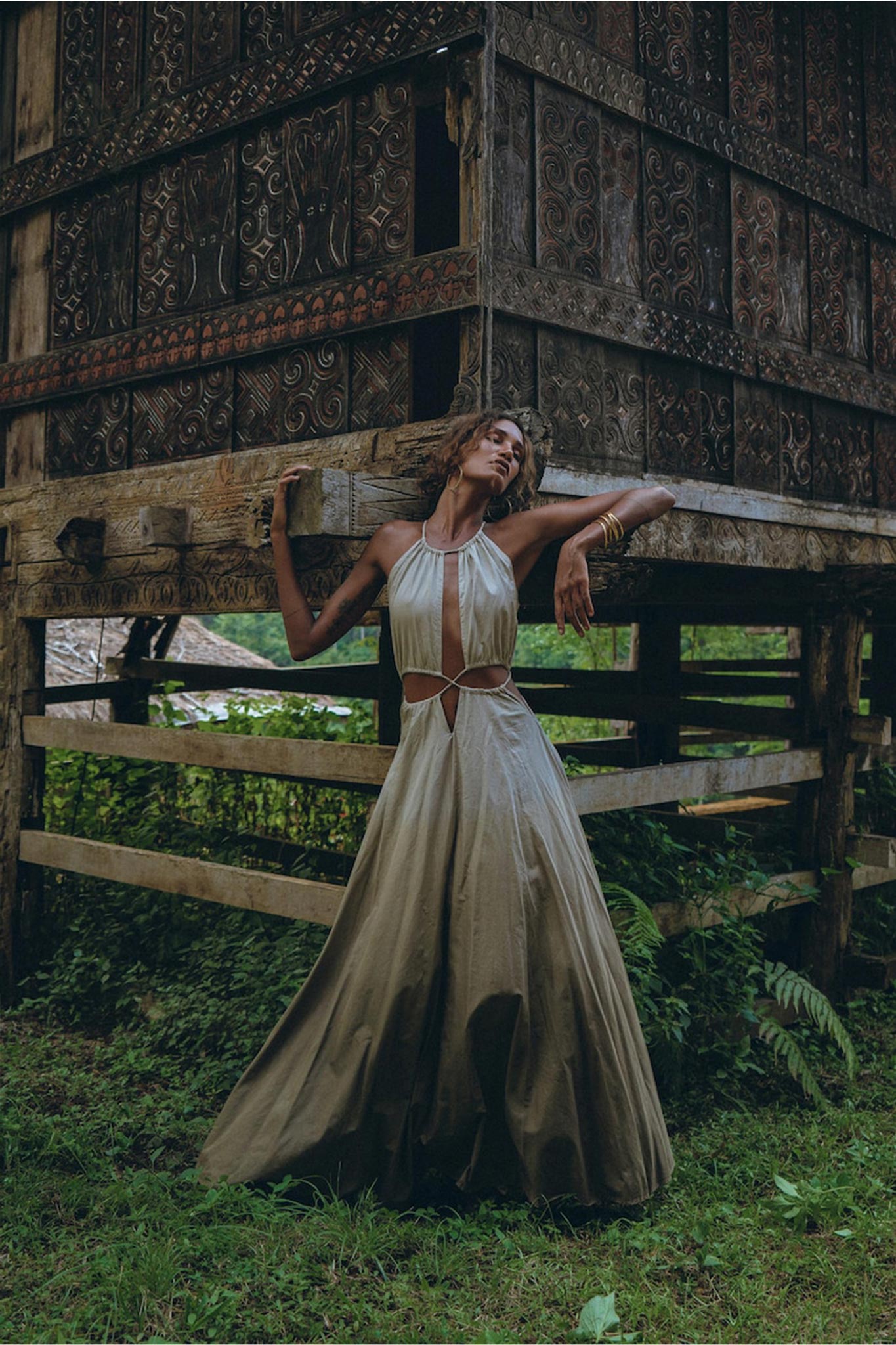 Wearing the Boho Bridesmaid Dress for Women from AYA Sacred Wear, a woman stands under a rustic wooden structure with intricate carvings. The flowing beige dress, crafted from handwoven fabrics, complements the lush green background that enhances the natural ambiance. She leans back with one arm raised, highlighting her gold bangles.