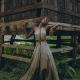 Wearing the Boho Bridesmaid Dress for Women from AYA Sacred Wear, a woman stands under a rustic wooden structure with intricate carvings. The flowing beige dress, crafted from handwoven fabrics, complements the lush green background that enhances the natural ambiance. She leans back with one arm raised, highlighting her gold bangles.