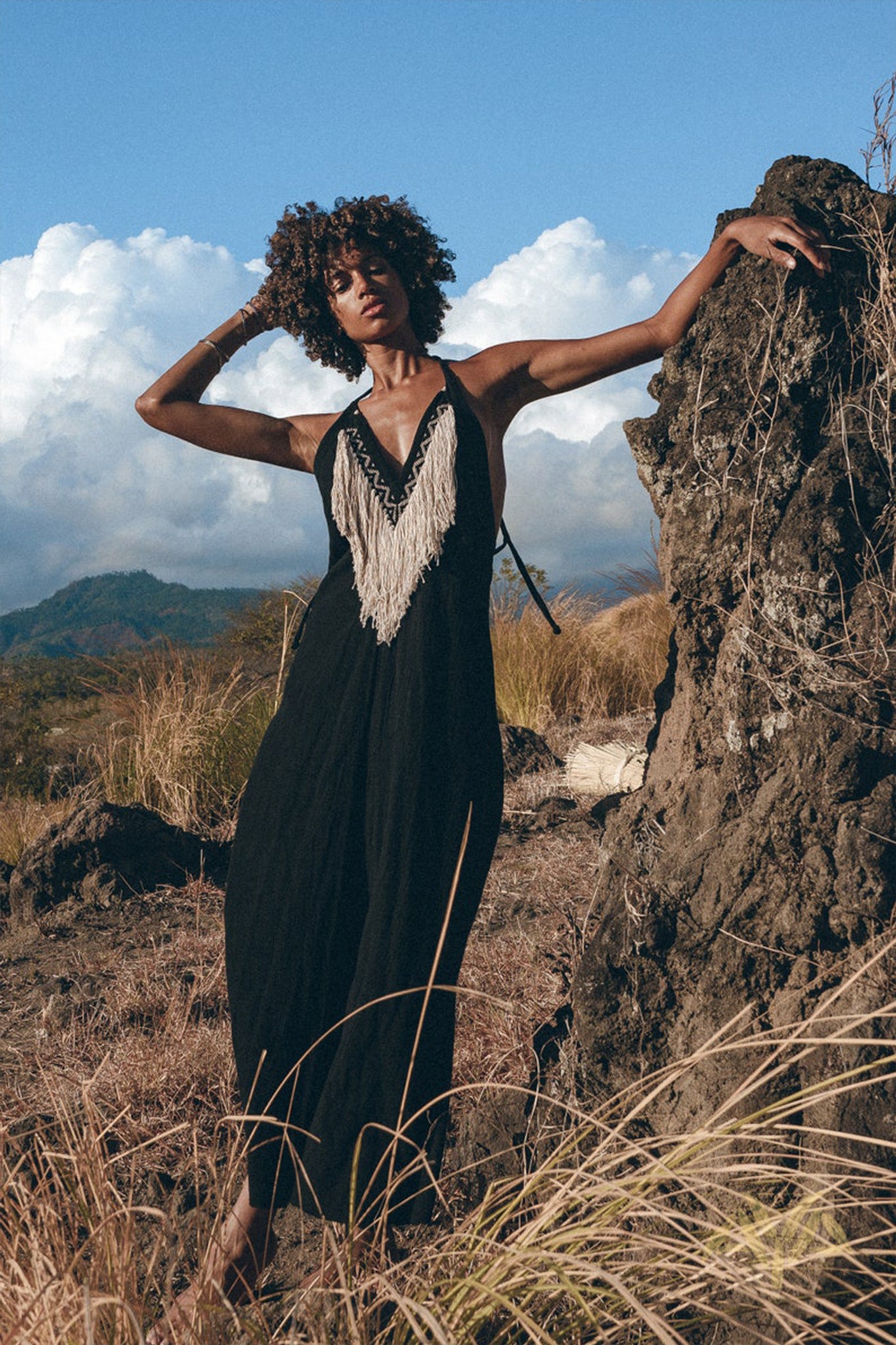 A person dressed in a long black Boho Dress by AYA Sacred Wear, featuring fringed tribal details, is posing outdoors while leaning against a large rock. The backdrop showcases dry grass, rocky terrain, and a partly cloudy blue sky.