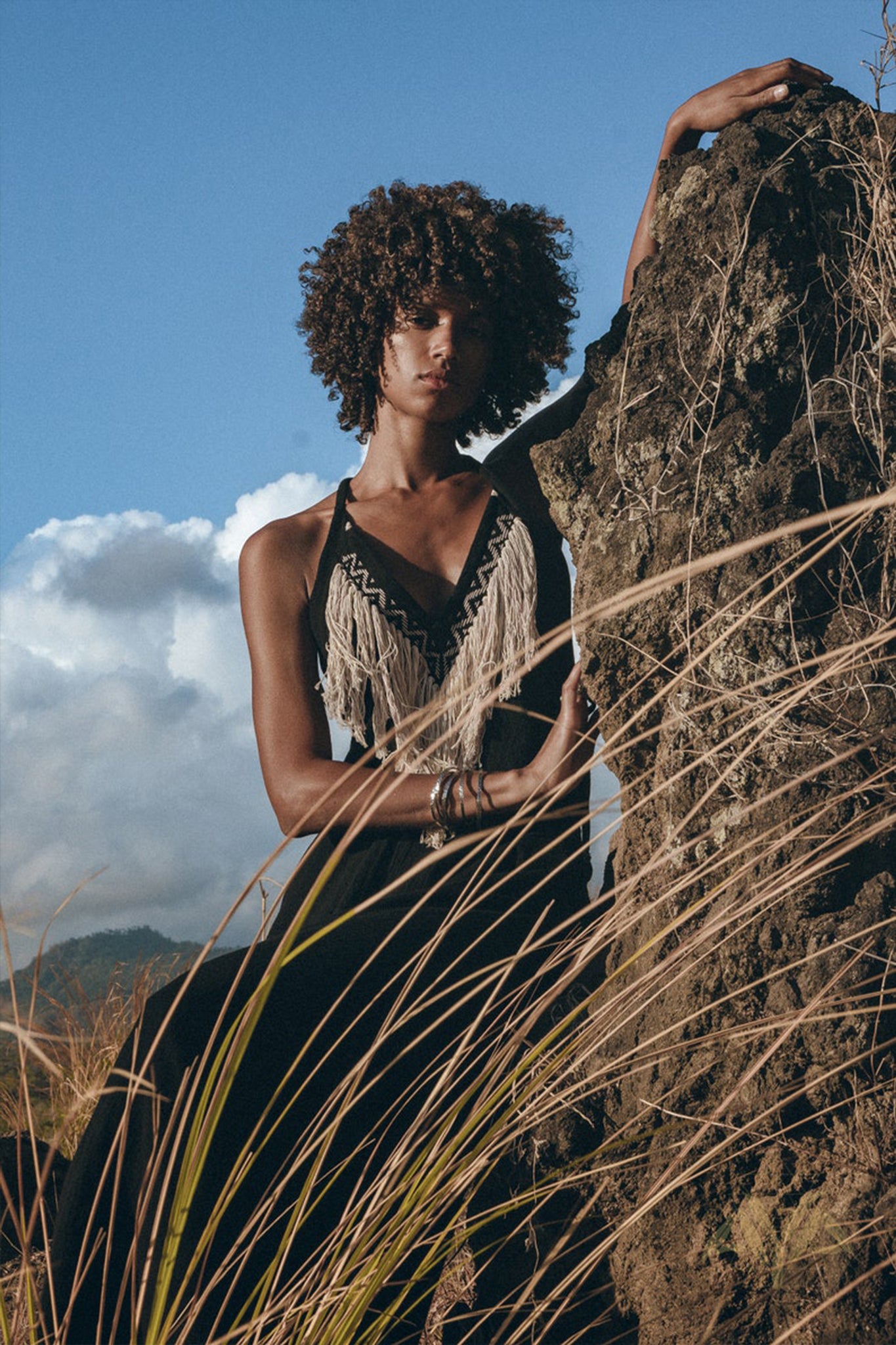 A woman with curly hair in an organic Bohemian Sleeveless Party Dress by AYA Sacred Wear, adorned with fringes, stands outdoors, leaning against a large rock. The natural setting, complete with tall grass and a clear blue sky, enhances the bohemian vibe of her elegant attire.