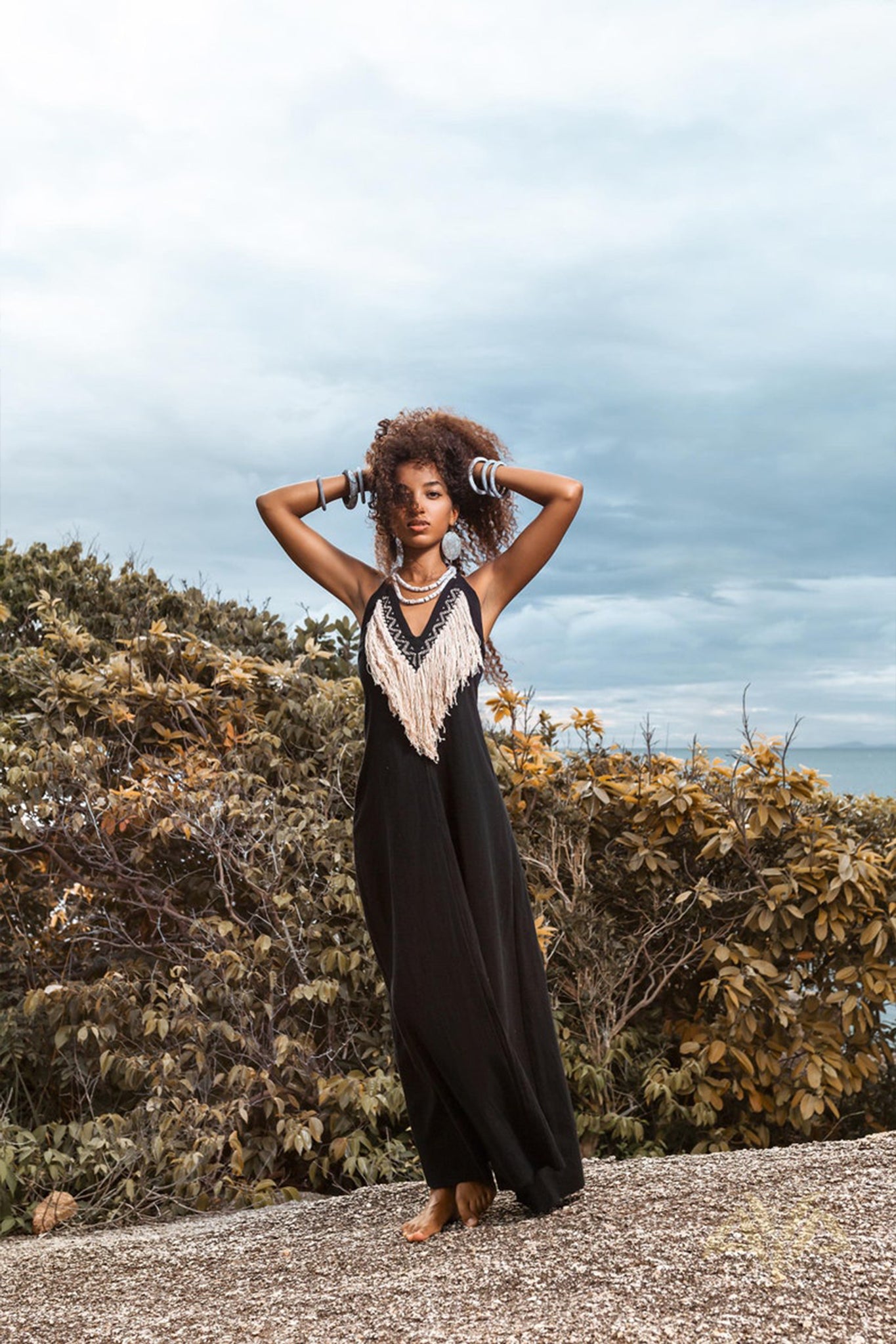 A person with curly hair poses confidently outdoors, wearing an AYA Sacred Wear black Boho Dress, featuring a fringed front detail. They stand on a stone surface, surrounded by trees under an overcast sky.