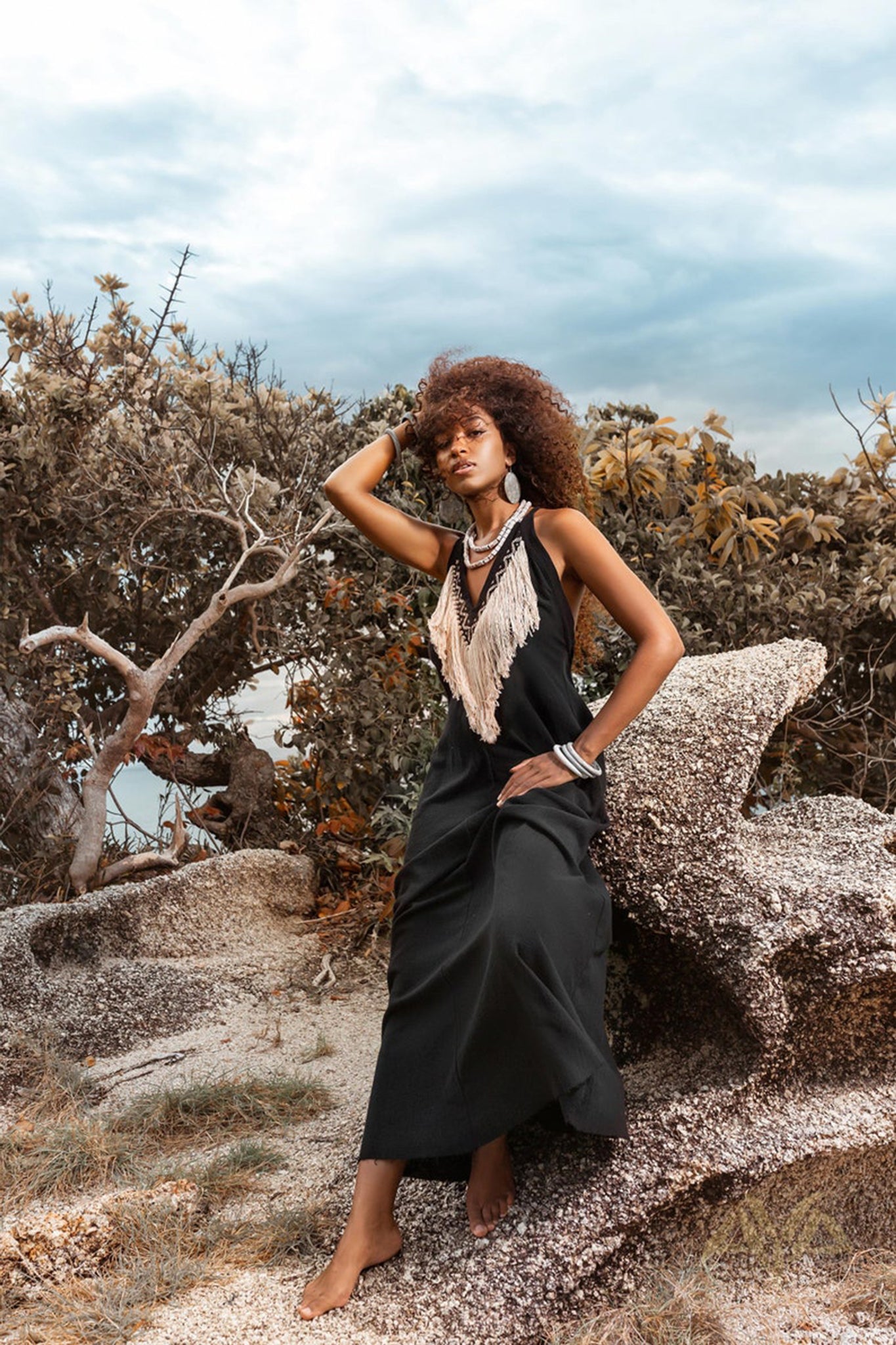 A woman with curly hair poses barefoot on large rocks wearing the Boho Dress by AYA Sacred Wear. This elegant organic bohemian, sleeveless party dress features fringed details and a striking tribal design. She stands amidst windswept foliage under a cloudy sky, accessorized with large earrings and bangles.