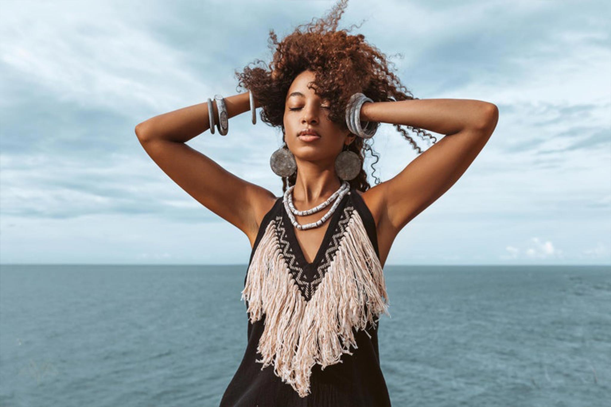 A woman with curly hair stands near the ocean, eyes closed and arms raised, wearing an organic bohemian dress from AYA Sacred Wear that captures a boho flair. Her look is further enhanced with silver jewelry, set against a backdrop of a cloudy sky and vast waters.