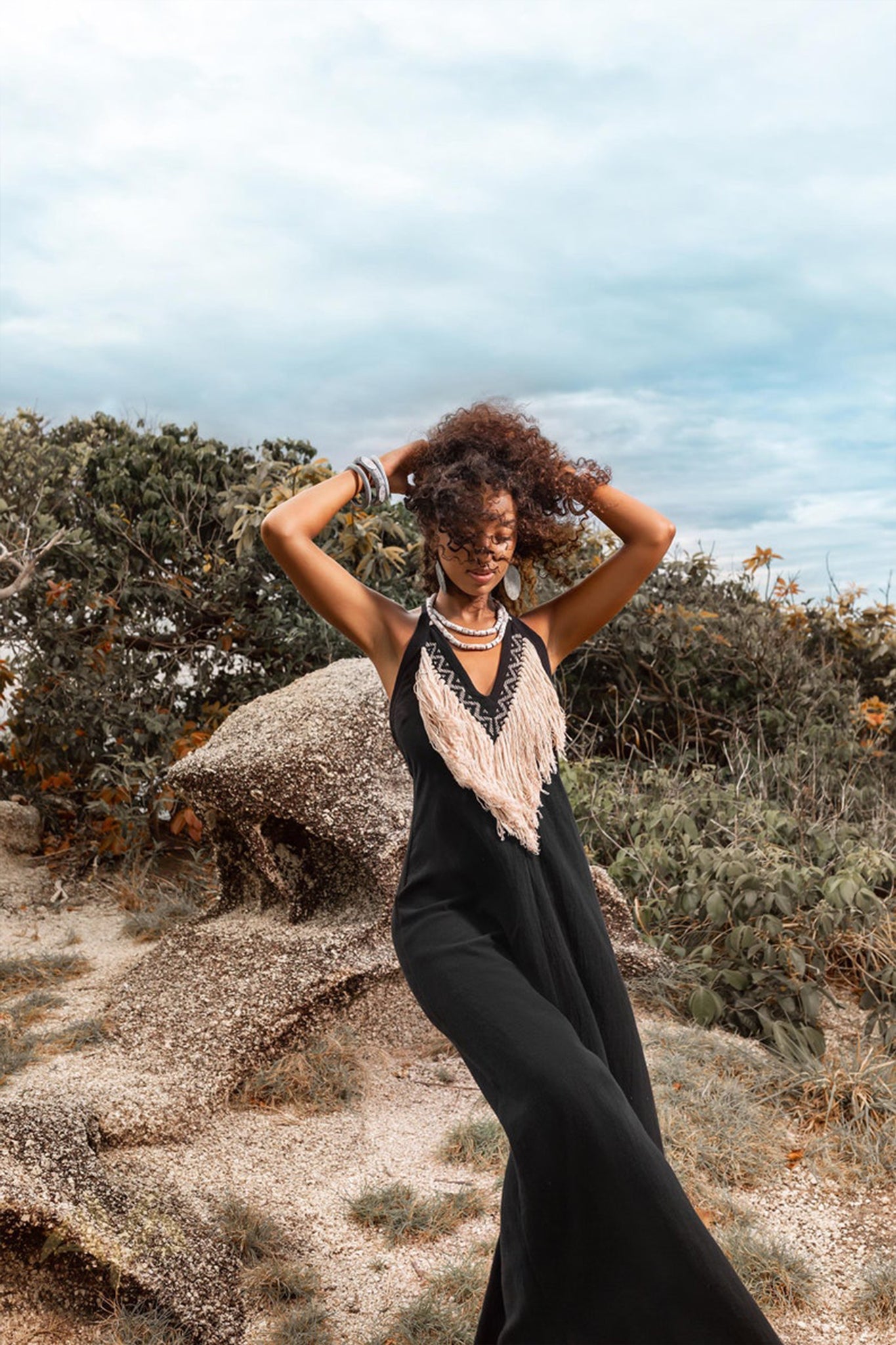 A person with curly hair stands outdoors on a rocky terrain, draped in an AYA Sacred Wear Organic Bohemian Dress featuring frayed details and complemented by jewelry. Surrounded by greenery under a cloudy sky, they hold their hair with both hands, creating a scene brimming with dynamic energy.