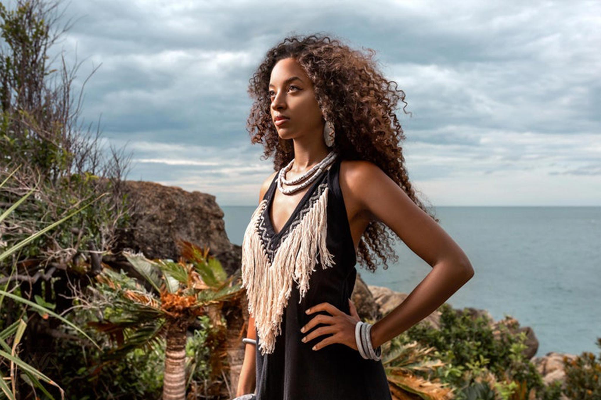 A woman with curly hair stands confidently outdoors near the ocean, beautifully dressed in a fringed top, adorned with necklaces and bracelets. She's wearing an organic bohemian dress from AYA Sacred Wear's collection, perfect for any event. The background showcases rocky terrain, lush greenery, and a cloudy sky.