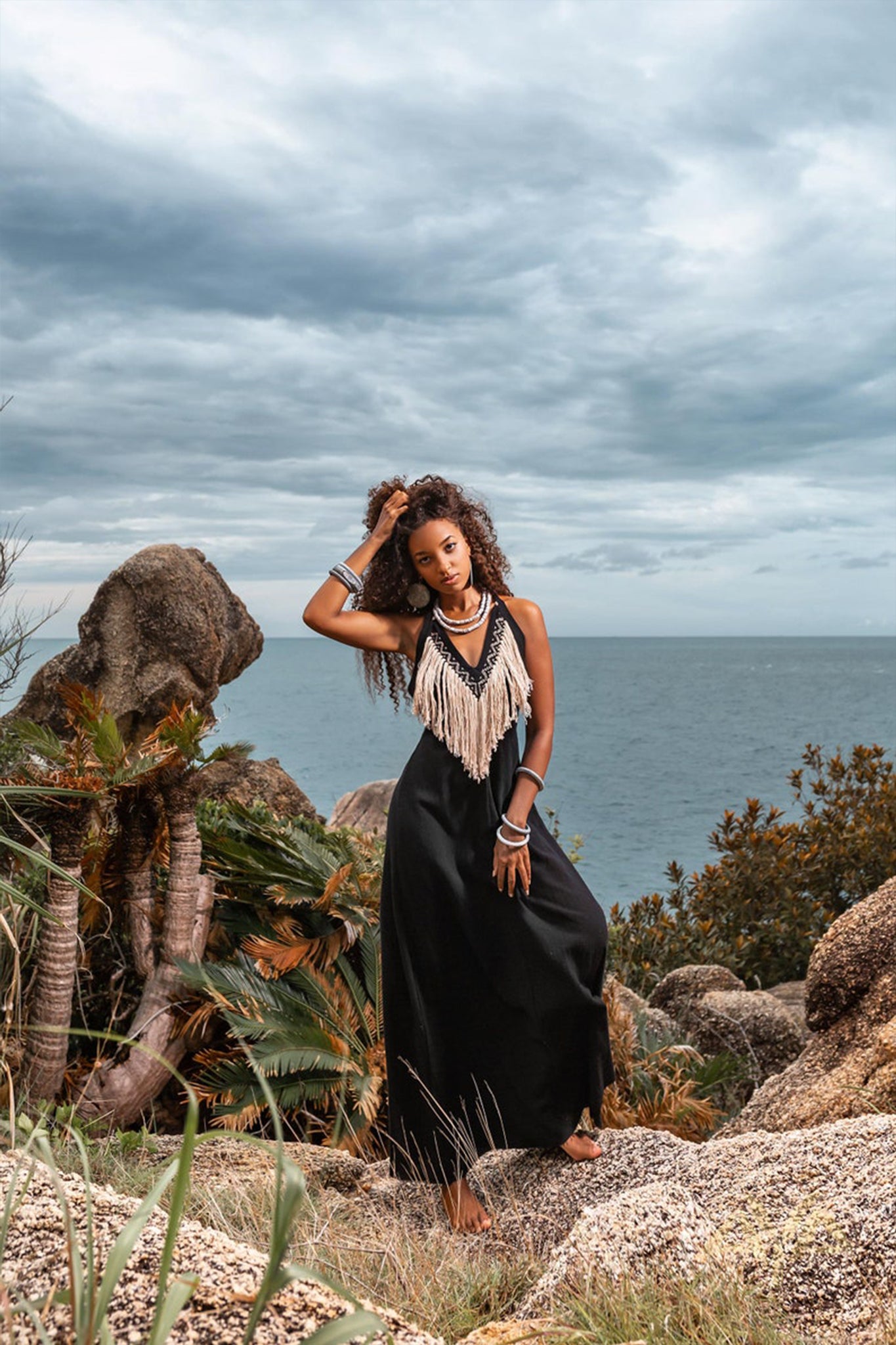 A woman in an AYA Sacred Wear flowing black Boho Dress, featuring fringe detail, poses barefoot on a rocky coastline. Surrounded by tropical plants and set against the backdrop of the ocean and a cloudy sky, she perfectly captures the essence of an organic bohemian scene.