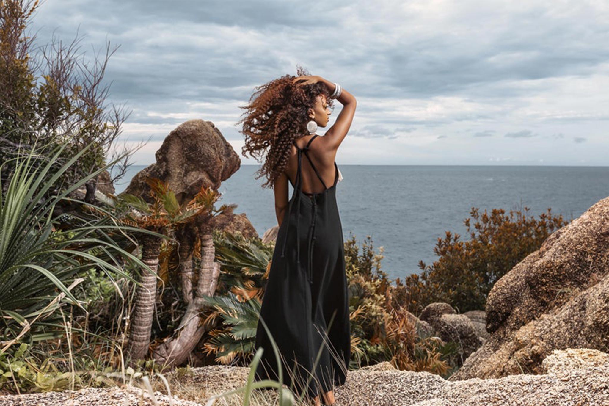 Draped in an AYA Sacred Wear Boho Dress, a woman stands atop a rocky hillside overlooking the ocean. Her curly hair swirls in the wind as she takes in the natural beauty surrounding her, with rocks and plants framing her against a backdrop of clouds stretching over the water.