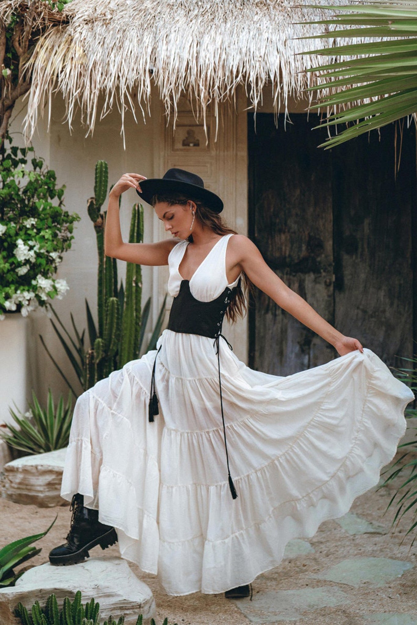 A woman in a wide-brimmed hat poses gracefully outdoors near a thatched-roof structure and cacti, wearing the Off-White Maxi Adjustable Bohemian Dress by AYA Sacred Wear. She pairs it with black boots and holds the dress's hem, showcasing its movement and design.