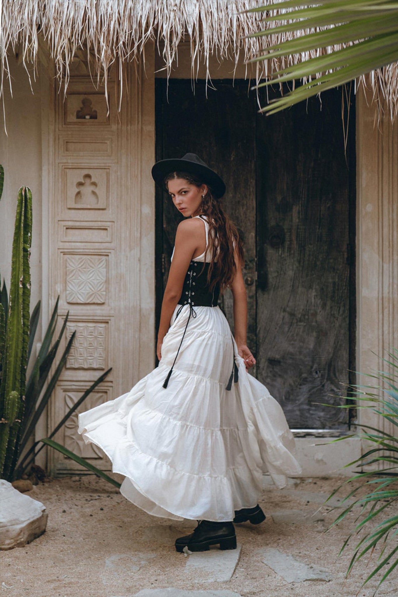 A woman wearing an AYA Sacred Wear Boho Dress for Women - an off-white maxi adjustable dress - stands in front of an ornate wooden door. Dressed in a black hat and black top, she looks back over her shoulder, enveloped by cacti and palm leaves in a rustic outdoor setting.