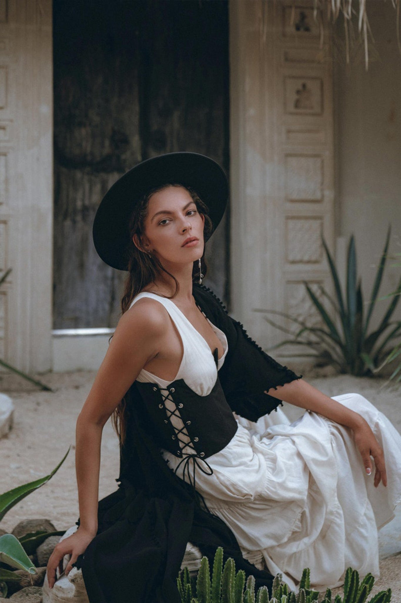 A woman in an AYA Sacred Wear Boho Dress for Women, an off-white maxi dress with adjustable features, sits outdoors. Her bohemian outfit is enhanced by a black corset and a wide-brimmed black hat, along with a sheer shawl. She is surrounded by desert plants, with a rustic wooden door providing the perfect backdrop for her eclectic style.