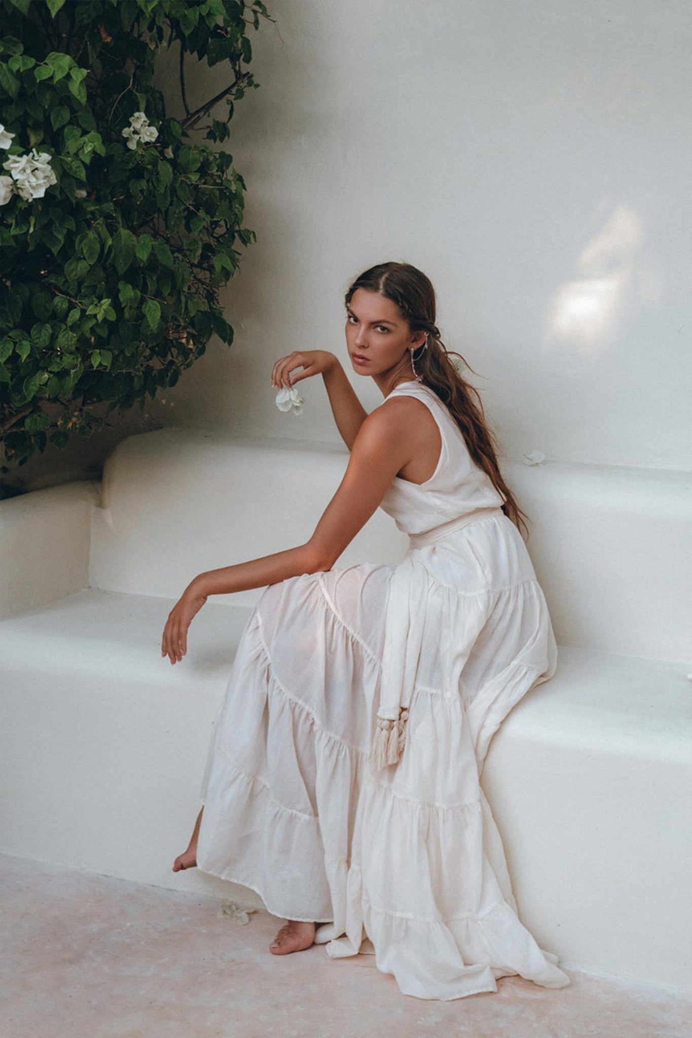 A woman dressed in an off-white Boho Dress by AYA Sacred Wear sits on a white bench, holding a small flower. She gazes thoughtfully to the side, surrounded by lush green plants in a serene setting.