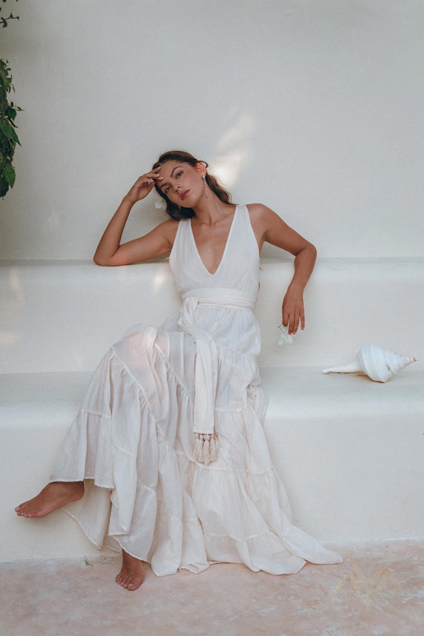 An elegant woman, wearing the Off-White Maxi Adjustable Dress from AYA Sacred Wear's Bohemian collection, sits gracefully on a minimalist white bench. Her head rests thoughtfully on her hand as she gazes contemplatively into the distance. Beside her lies a large white seashell, while green foliage nearby enhances the scene's tranquil charm and organic cotton elegance.