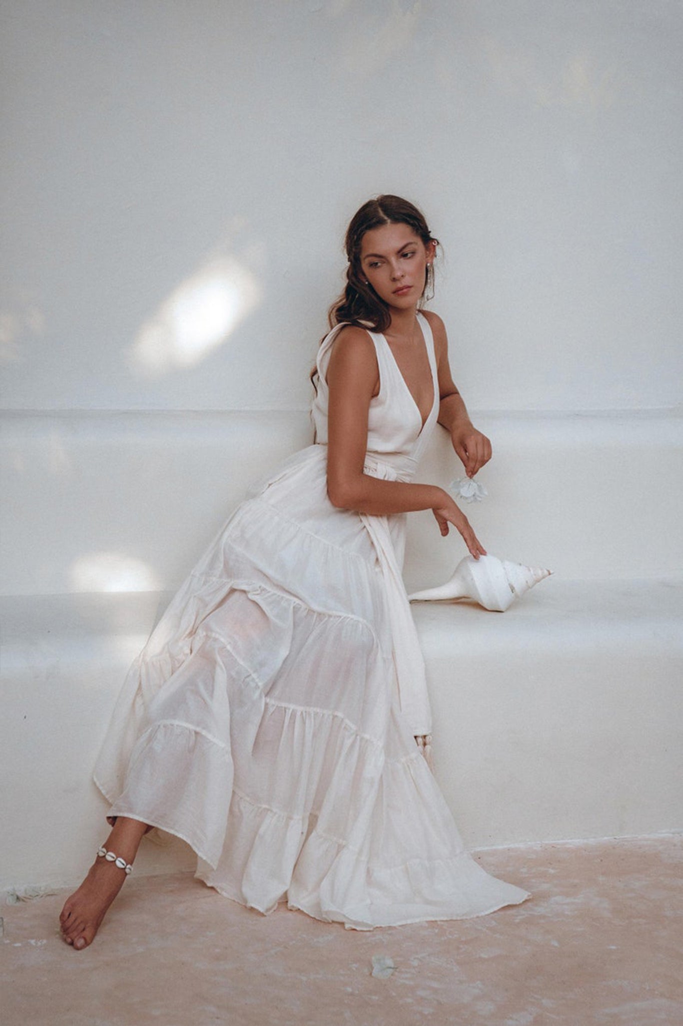 A woman in an off-white maxi adjustable dress from AYA Sacred Wear's Boho Dress for Women collection sits barefoot on a minimalist white bench. She gazes thoughtfully to the side, holding a large seashell. Soft lighting creates a serene and dreamy atmosphere.