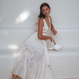A woman in an off-white maxi adjustable dress from AYA Sacred Wear's Boho Dress for Women collection sits barefoot on a minimalist white bench. She gazes thoughtfully to the side, holding a large seashell. Soft lighting creates a serene and dreamy atmosphere.