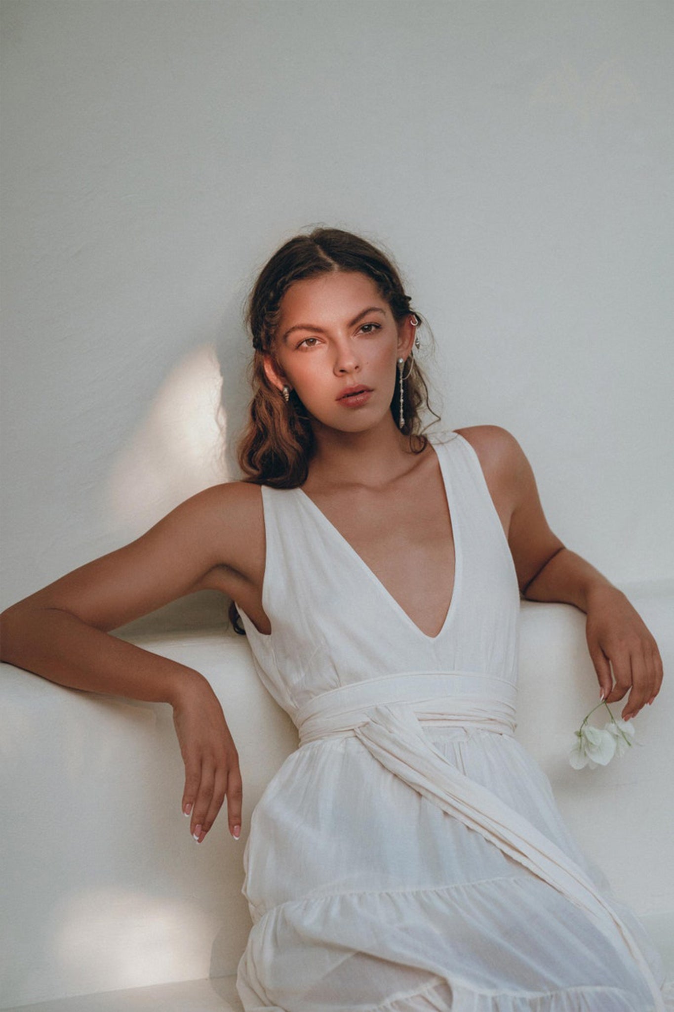 An elegant woman with long hair sits gracefully, adorned in a Boho Dress for Women by AYA Sacred Wear. This off-white maxi adjustable dress features a sleeveless design with a deep V-neck, perfectly complementing the white rose she holds. Soft lighting creates gentle shadows in the background, enhancing her ethereal elegance.