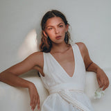 An elegant woman with long hair sits gracefully, adorned in a Boho Dress for Women by AYA Sacred Wear. This off-white maxi adjustable dress features a sleeveless design with a deep V-neck, perfectly complementing the white rose she holds. Soft lighting creates gentle shadows in the background, enhancing her ethereal elegance.