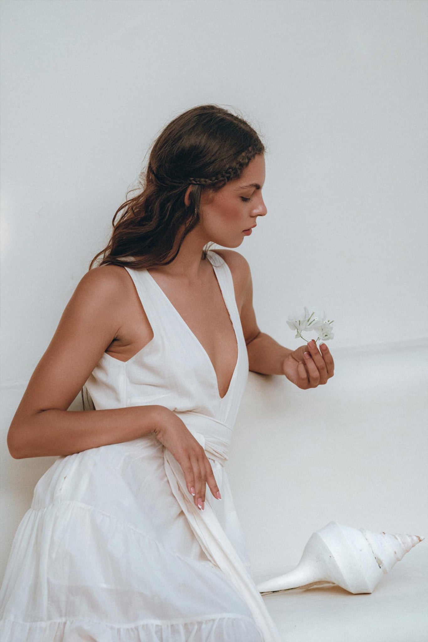 A woman dressed in AYA Sacred Wear's Boho Dress for Women in off-white sits pensively against a light background, holding a small flower. Beside her rests a large seashell, enhancing the scene's serene and minimalistic vibe.