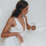 A woman dressed in AYA Sacred Wear's Boho Dress for Women in off-white sits pensively against a light background, holding a small flower. Beside her rests a large seashell, enhancing the scene's serene and minimalistic vibe.