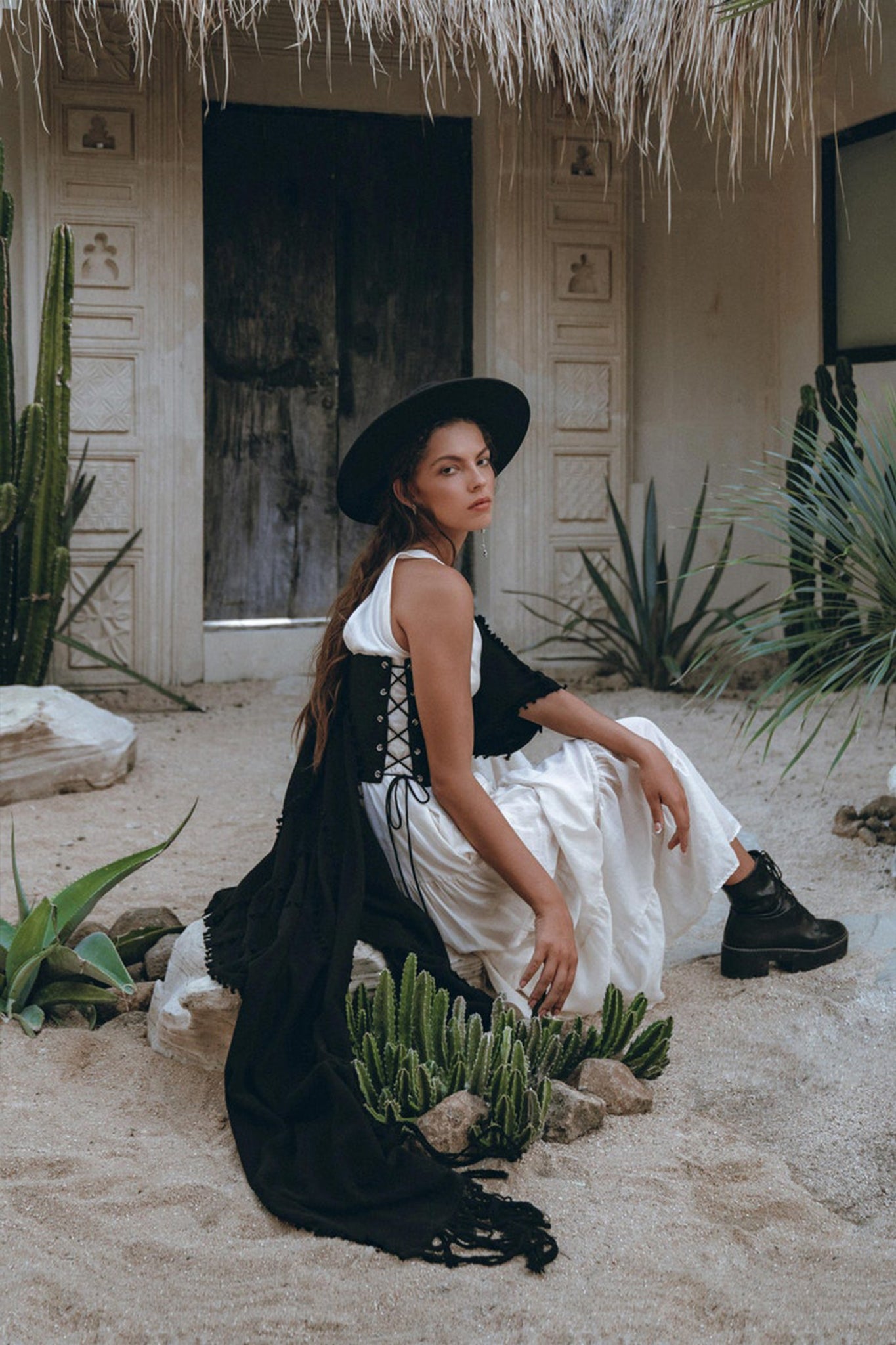 A woman in an off-white AYA Sacred Wear Boho Dress for Women, an adjustable maxi dress with a Bohemian flair, sits on sandy ground surrounded by succulents. She's accessorized with a wide-brimmed black hat and chunky black boots. In the background stands a rustic building with carved wooden doors.