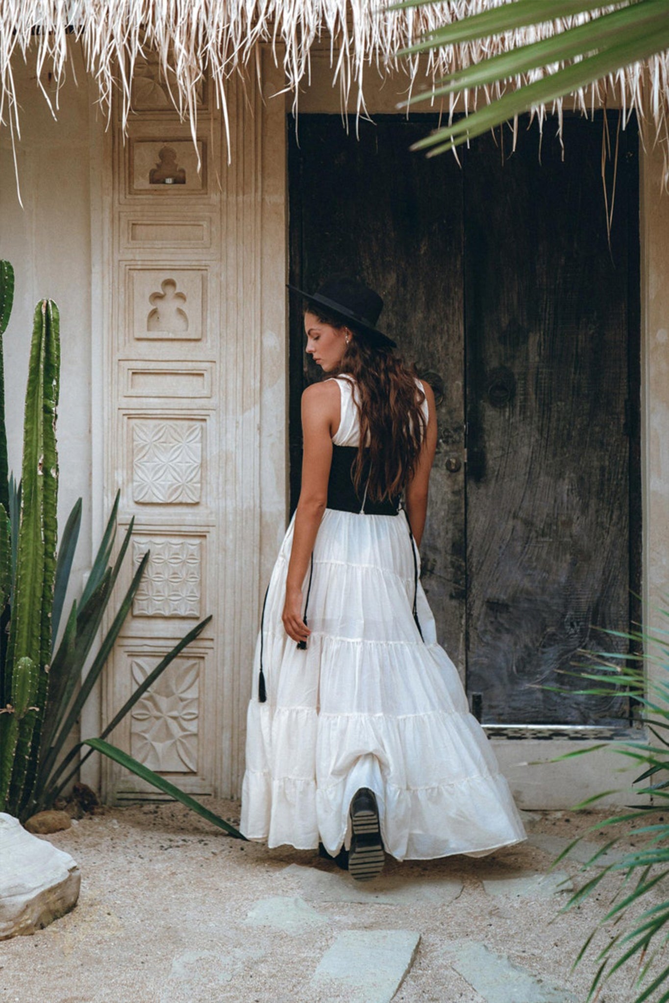 A woman in an off-white maxi Boho Dress by AYA Sacred Wear, complemented by a black hat, stands in front of a rustic wooden door amidst tall green cacti and textured walls under a thatched roof.