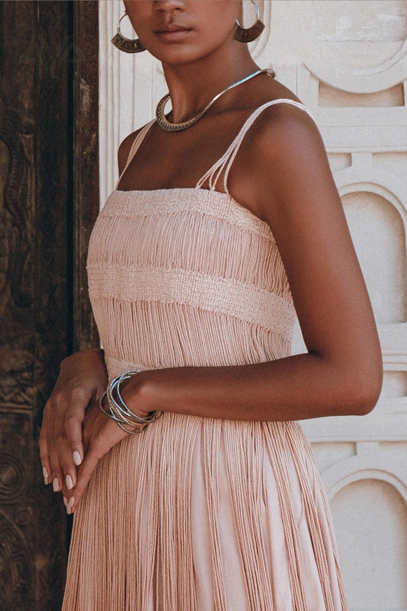 A woman poses with her hands crossed in front, wearing an Ethnic Handwoven Macrame Dress by AYA Sacred Wear. The sleeveless dress in dusty pink is crafted from organic cotton and features a tribal design. Her look is completed with silver hoop earrings, a necklace, and bangles. The setting emphasizes an intricate architectural wall while her face remains hidden from view.