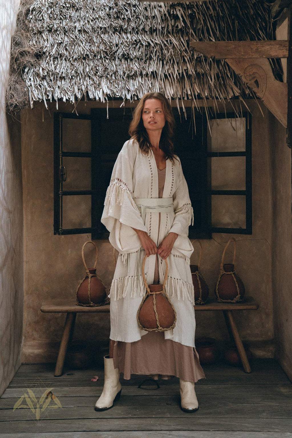 In a rustic setting with a thatched roof and wooden bench, an individual is seen wearing the Boho Kimono Cardigan by AYA Sacred Wear. This layered, textured robe harmonizes traditional and contemporary elements as they hold a decorative jug and sport white boots.